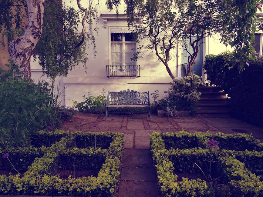 A small, well-maintained garden featuring a paved patio area with a decorative wrought iron bench placed against a white exterior wall of a house. The garden is bordered by neatly trimmed boxwood hedg